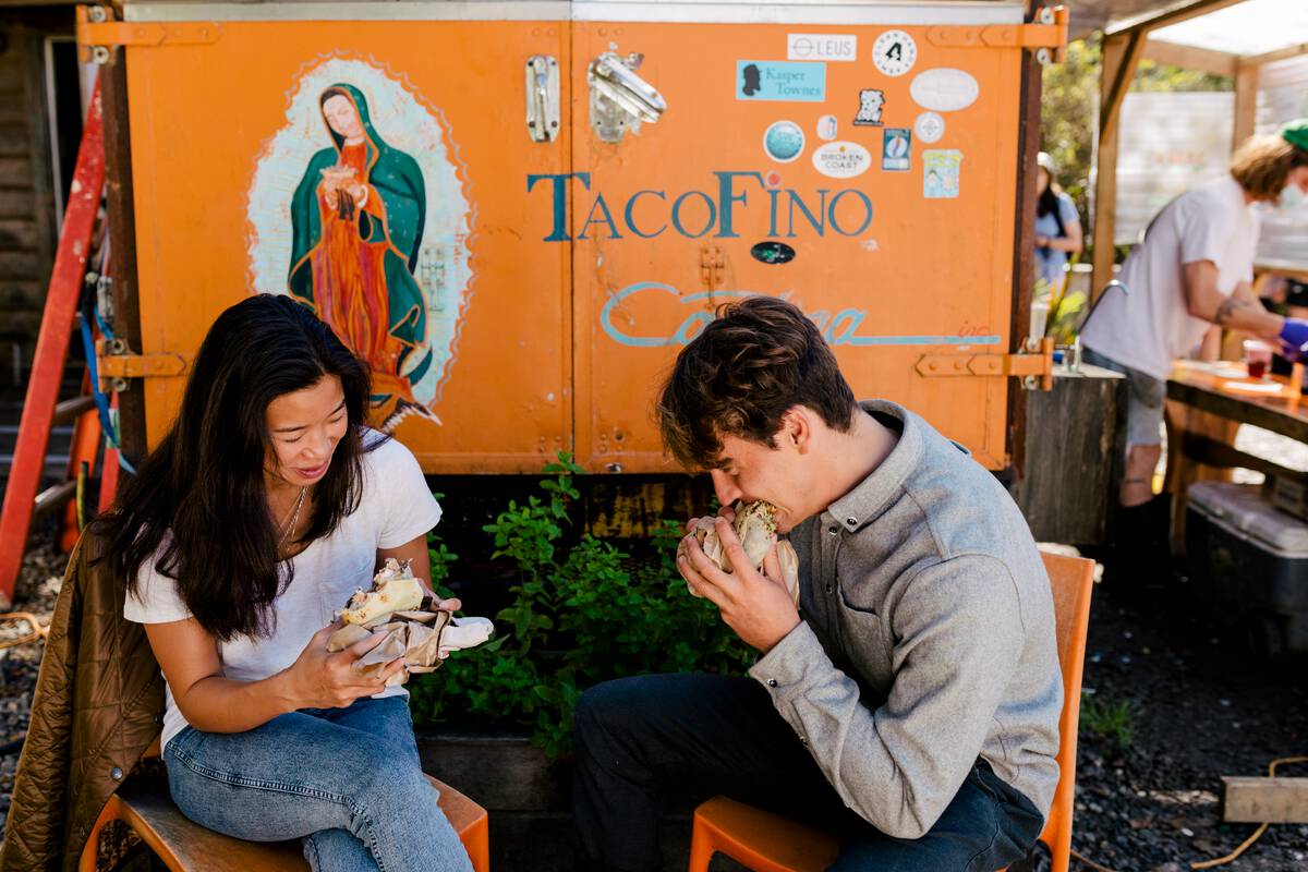 Two people each burritos behind then Tacofino food truck. They sit on orange chairs on a sunny day.