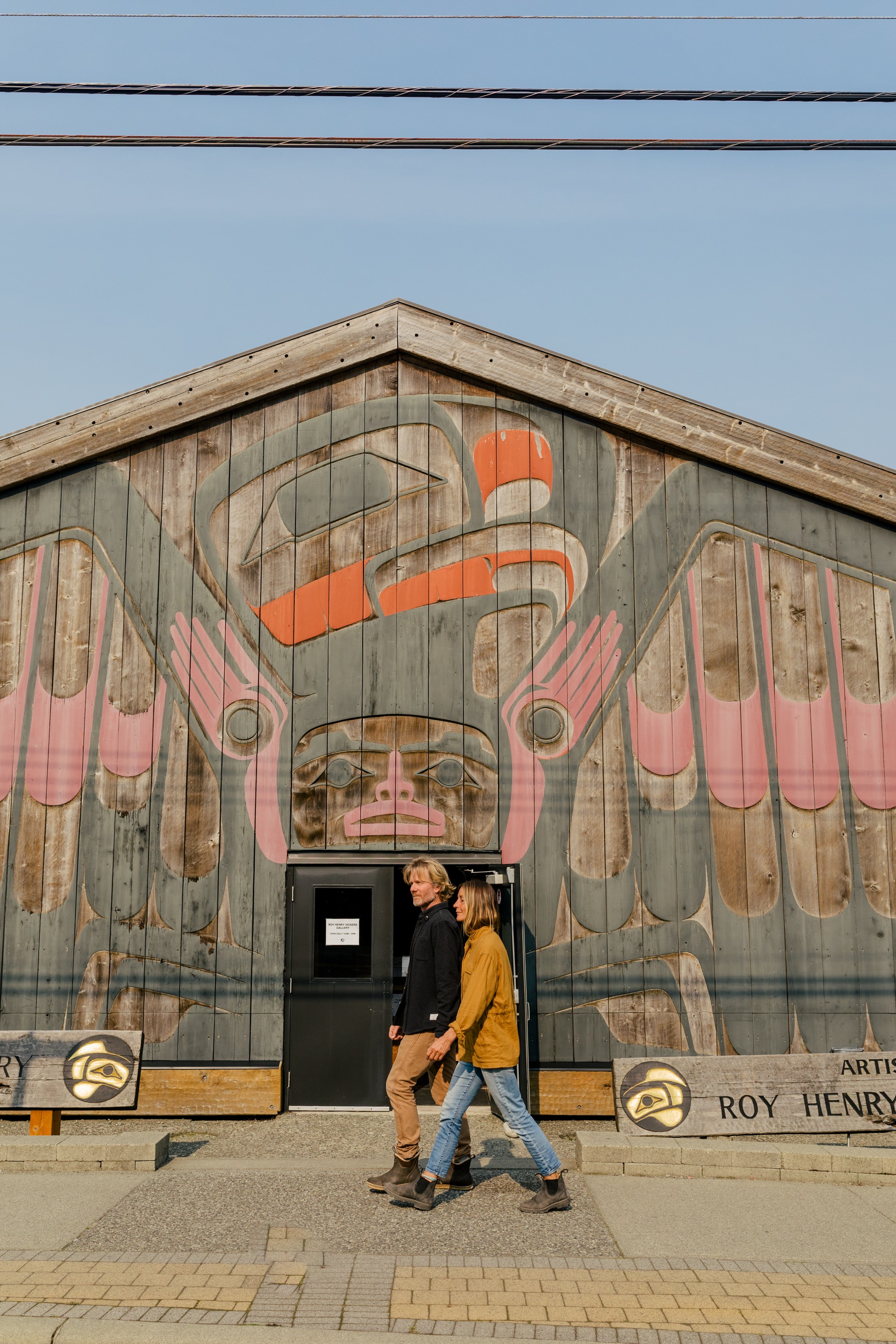 A couple walks out front of the Roy Henry Vickers Gallery on a clear sky day. Indigenous artwork is painted onto the front of the gallery.